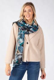 A woman modeling a beige textured cardigan and a blue and gray abstract print scarf, paired with blue jeans, against a plain white studio background.