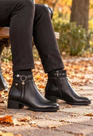 Black leather ankle boots with side zippers, low block heels, and a decorative silver ring and heart charm on the outer strap, shown on a person sitting on a bench outdoors in autumn.