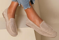 A pair of beige laser loafer flats worn by a person sitting with crossed legs, showing the soft squared toe slip-on design with a faux suede finish.