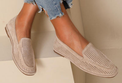 A pair of beige laser loafer flats worn by a person sitting with crossed legs, showing the soft squared toe slip-on design with a faux suede finish.