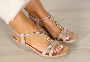A pair of silver and gold floral and pearl gem sandals with elastic ankle straps and a low wedge heel, worn by a person with light colored pants.