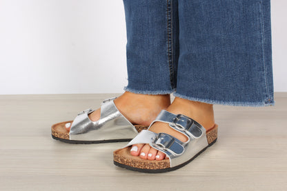 A foot wearing a double strap sandal with silver buckles, the sandal is silver, the buckle is silver, the foot is in a relaxed position.