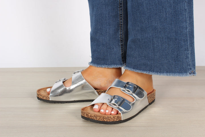 A foot wearing a double strap sandal with silver buckles, the sandal is silver, the buckle is silver, the foot is in a relaxed position.