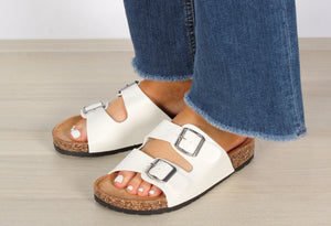A foot wearing a double strap sandal with silver buckles, the sandal is  white, the buckle is silver, the foot is in a relaxed position.