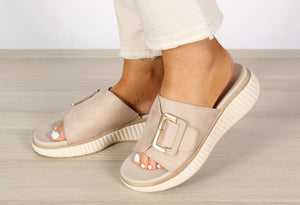 A pair of beige slip-on sandals with buckle detail and open toe, worn by a person standing on a light wooden floor.