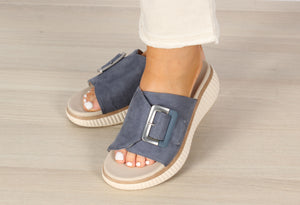 A pair of beige slip-on sandals with buckle detail and open toe, worn by a person standing on a light wooden floor.