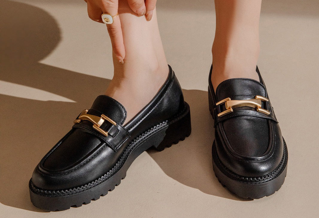 Black loafers with gold accents worn by a person on a beige background