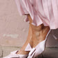 Pink high heels with bow details worn by a person in a pink skirt against a light-colored wall.