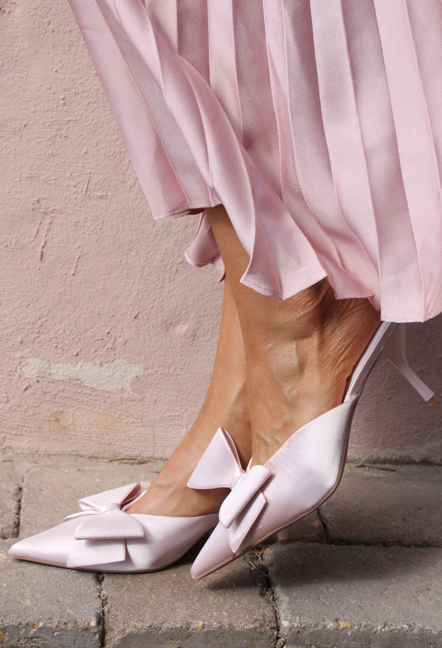 Pink high heels with bow details worn by a person in a pink skirt against a light-colored wall.