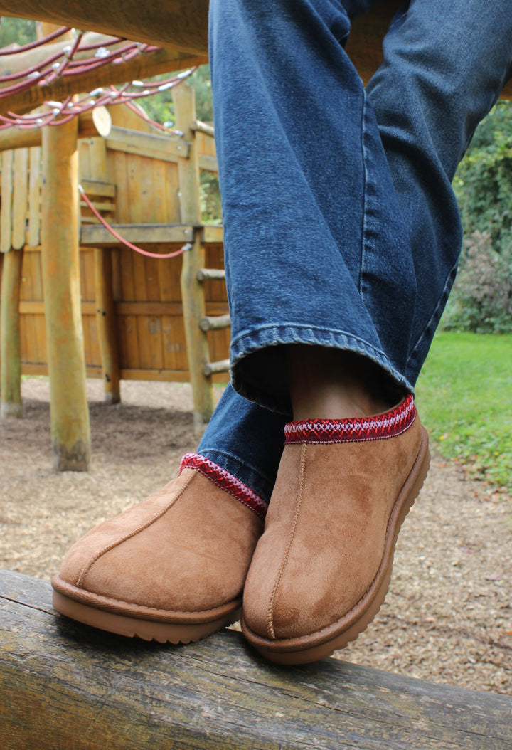 A person wearing slip-on boots with a round toe and fully faux fur lining, featuring embroidered details. The boots are primarily brown with red and beige accents.