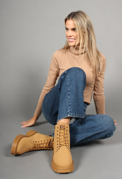 A woman sitting on a grey background wearing a beige turtleneck sweater, blue jeans, and camel 'Working Style Boots' with a round toe, lace-up front, faux fur lining, and a block heel.