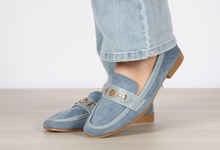 A person wearing blue faux suede loafers with leather look trims and antique gold hardware, the shoes are a slip-on style.