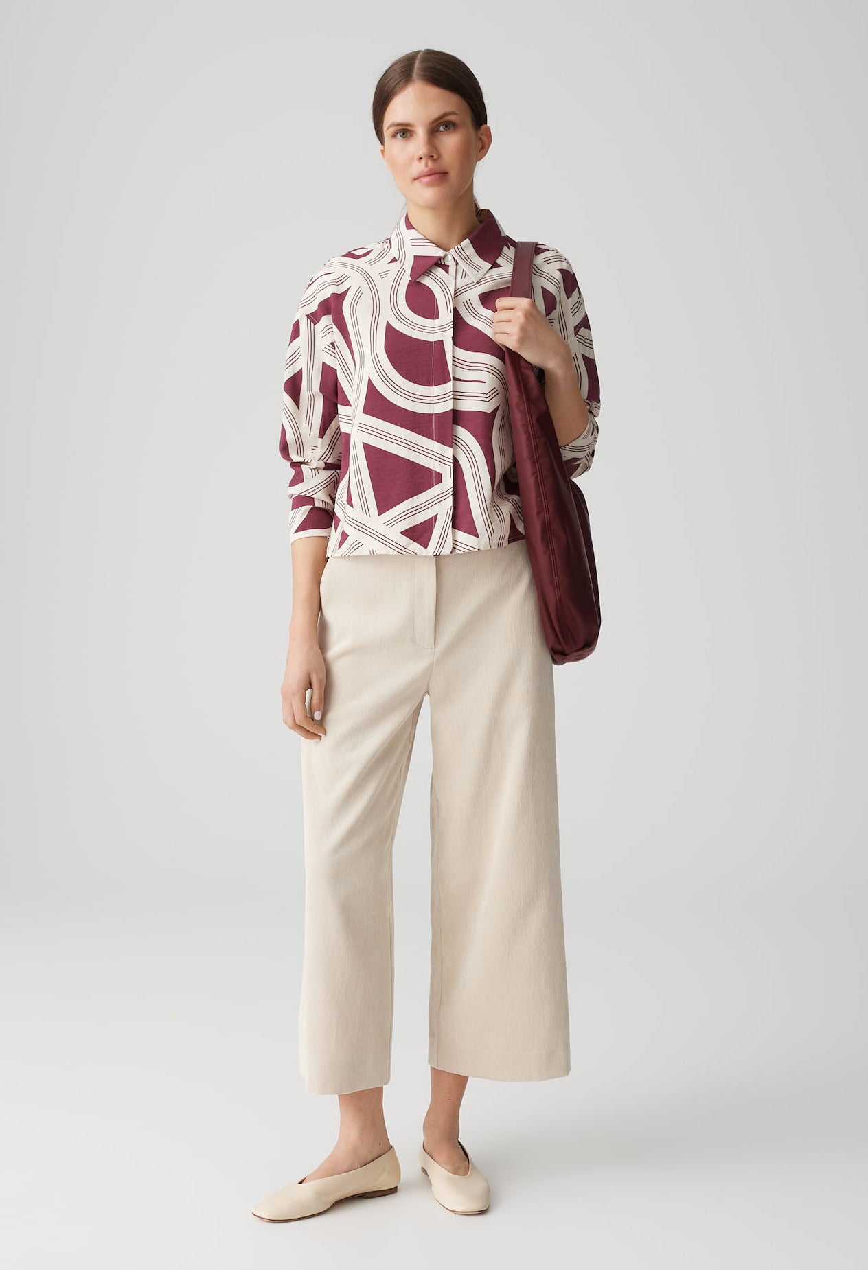 Woman wearing a patterned blouse and beige pants with a red bag on a white background