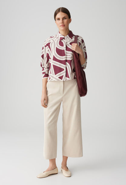 Woman wearing a patterned blouse and beige pants with a red bag on a white background