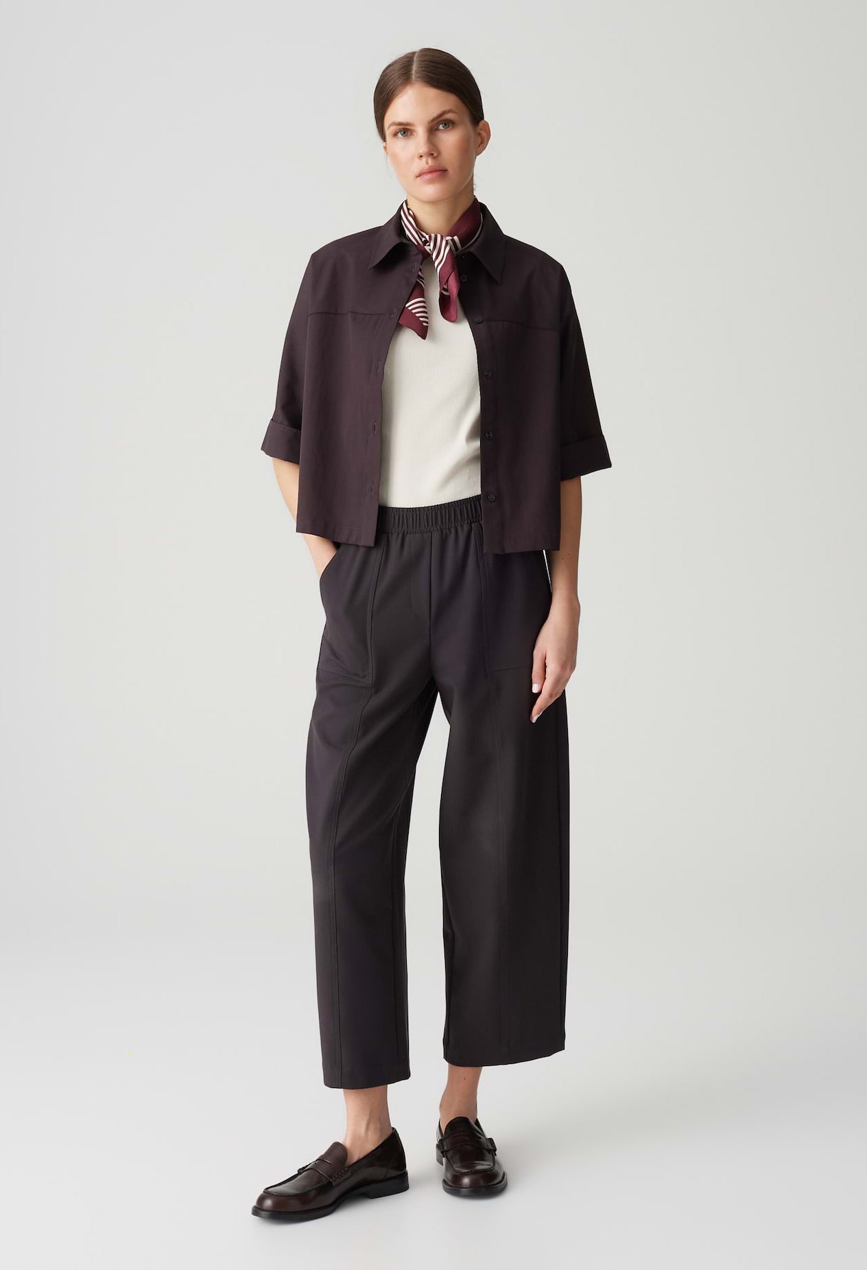 Woman wearing a dark brown jacket, white shirt, and black pants on a light gray background