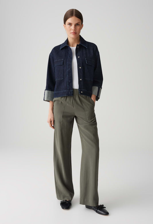 Person wearing a navy denim jacket, white shirt, and olive green pants on a light gray background