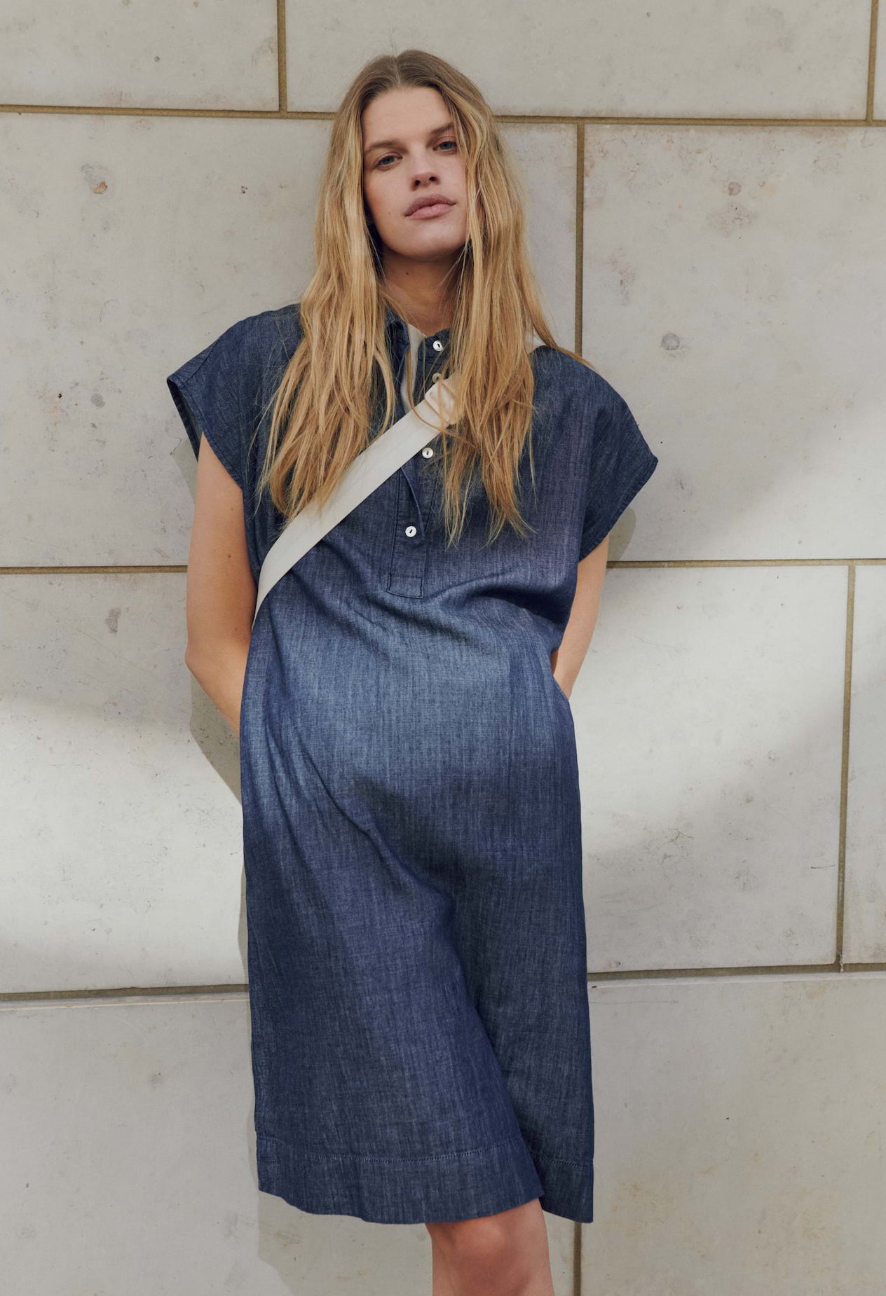 Woman wearing a blue dress with a beige strap over her shoulder against a tiled wall.
