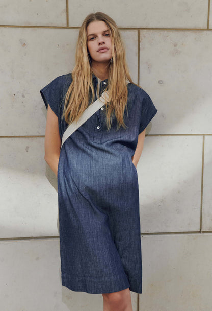 Woman wearing a blue dress with a beige strap over her shoulder against a tiled wall.