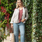 Woman standing against a wall of greenery wearing a beige jacket, pink shirt, blue jeans, and brown shoes.