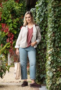 Woman standing against a wall of greenery wearing a beige jacket, pink shirt, blue jeans, and brown shoes.