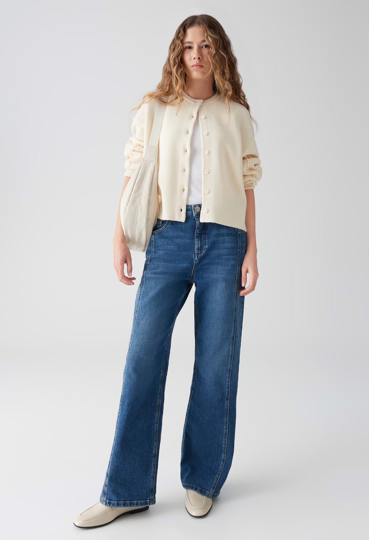 Woman wearing a cream blouse, white shirt, and blue jeans on a light gray background