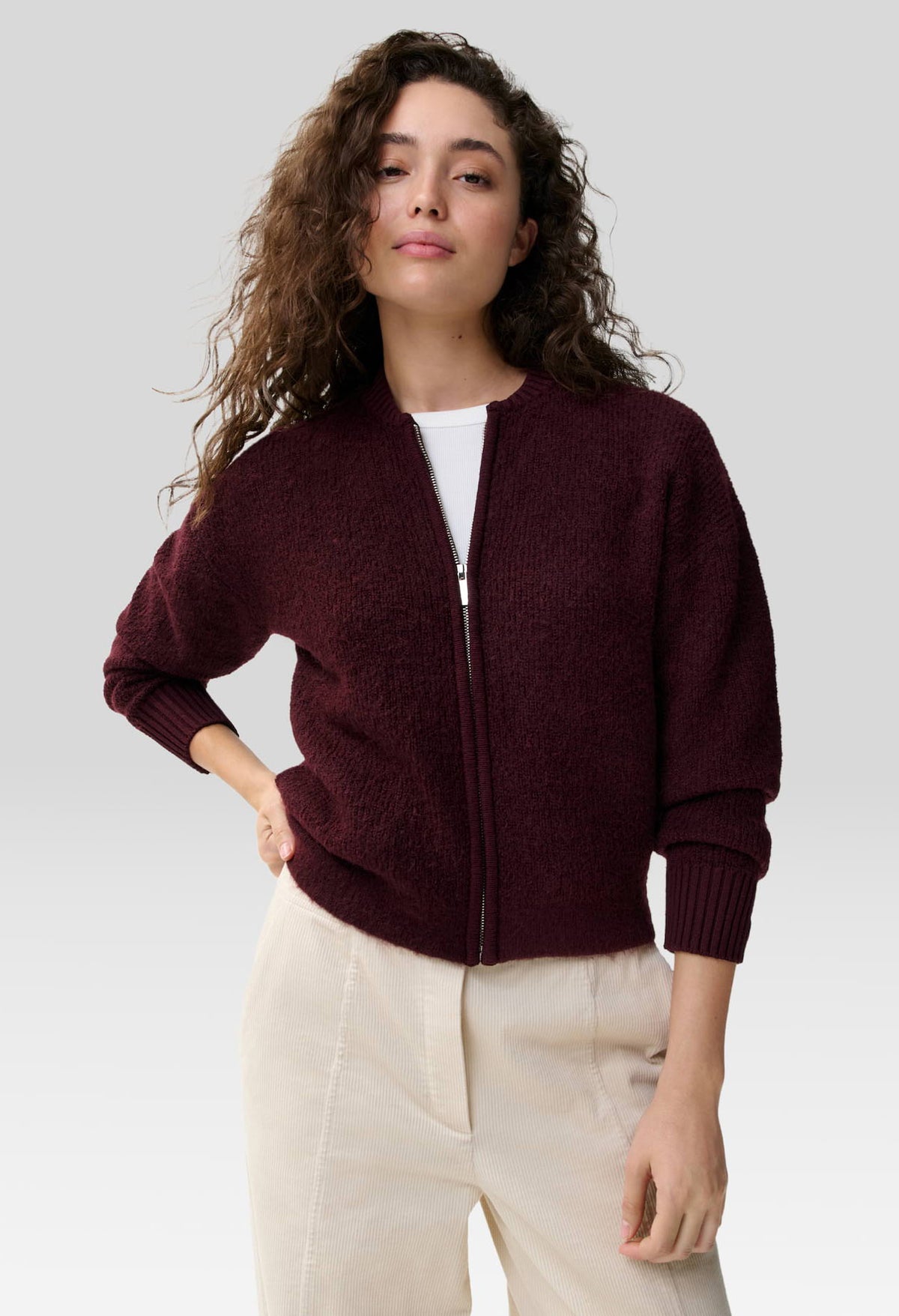 A woman wearing a textured, deep burgundy zip-up cardigan over a white top and cream-colored trousers, photographed against a neutral light gray studio background.