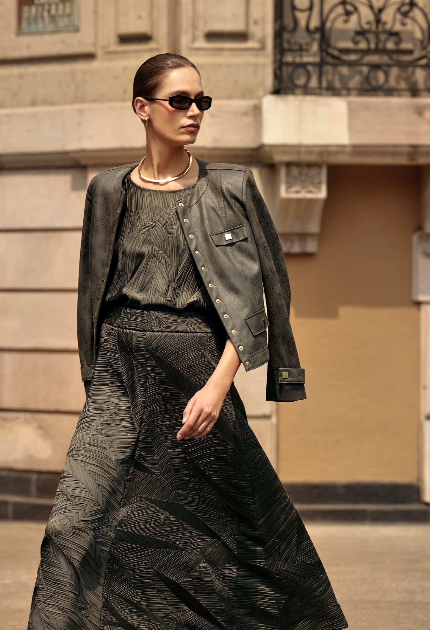 Woman in a stylish outfit with a jacket and sunglasses standing in front of a building.