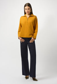 Woman wearing a mustard yellow sweater and dark blue pants on a white background