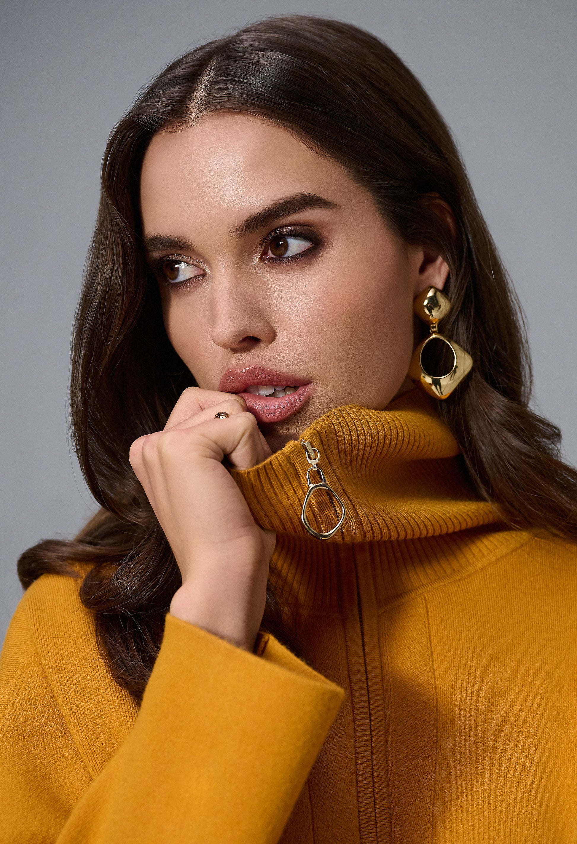 Woman wearing a mustard yellow coat with gold earrings against a gray background