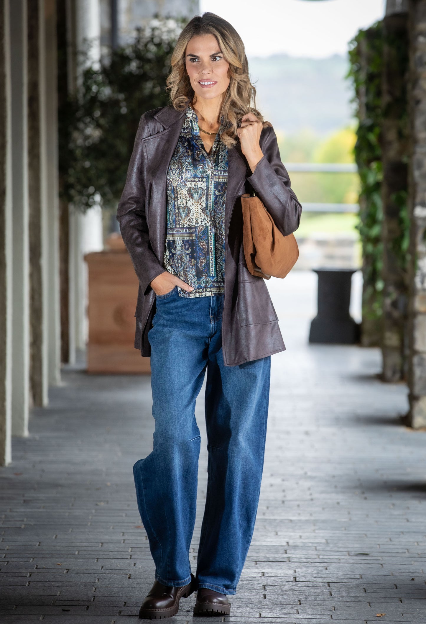 Woman in a stylish outfit with a jacket, patterned shirt, and jeans walking outdoors.