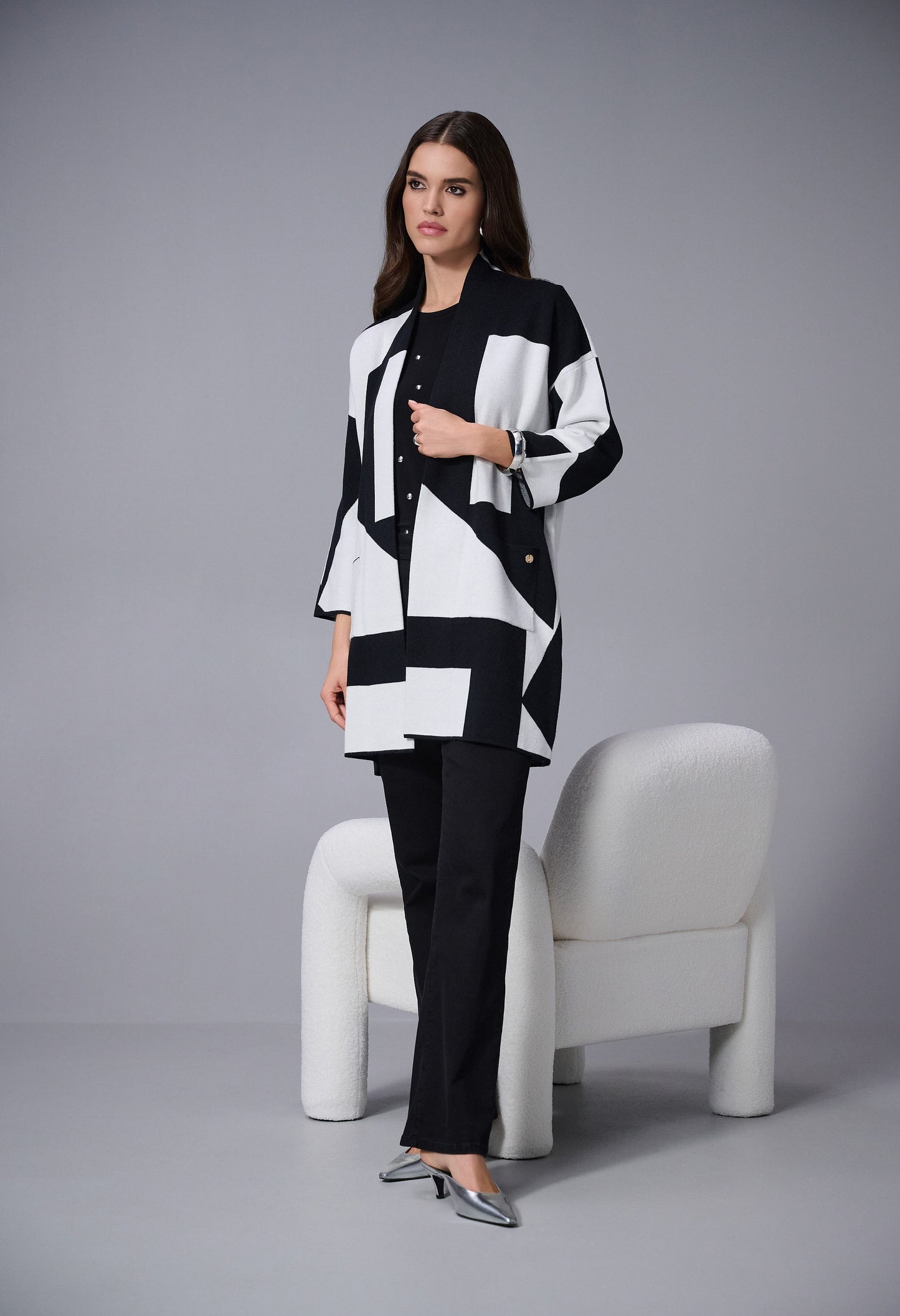 Woman wearing a black and white patterned outfit sitting on a white chair against a gray background