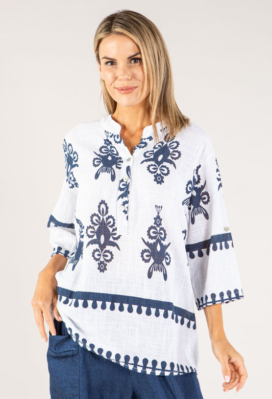 Woman wearing a white blouse with blue patterns and pom-pom details on a plain background