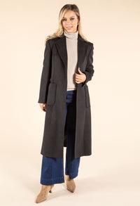 Woman wearing a long dark coat over a light sweater and jeans on a beige background