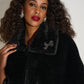 Woman wearing a black fur coat with a bow detail on a plain background
