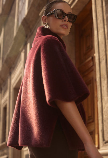 Woman wearing a burgundy cape and sunglasses in front of a stone building.
