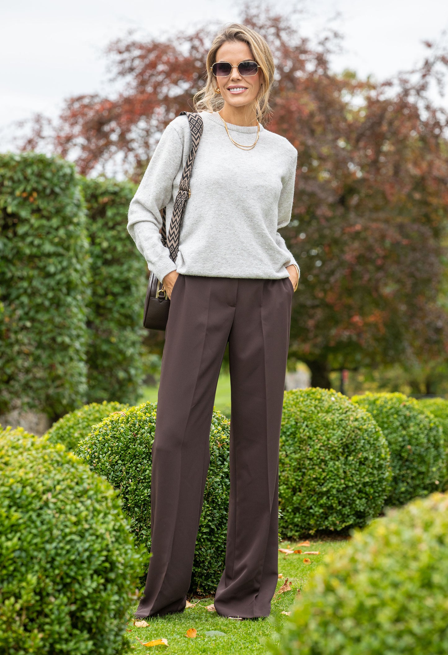 Woman in a light gray sweater and dark brown pants standing in a garden.