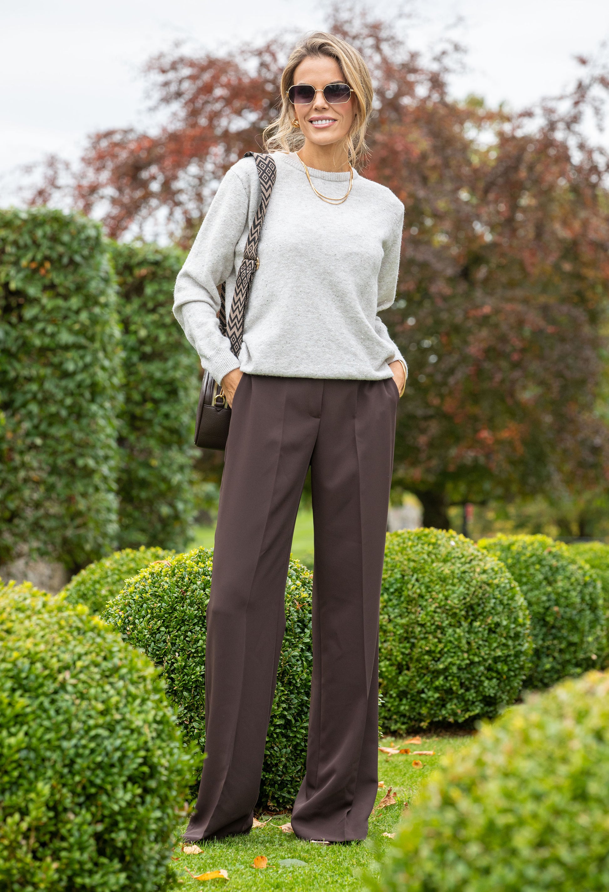 Woman in a light gray sweater and dark brown pants standing in a garden.