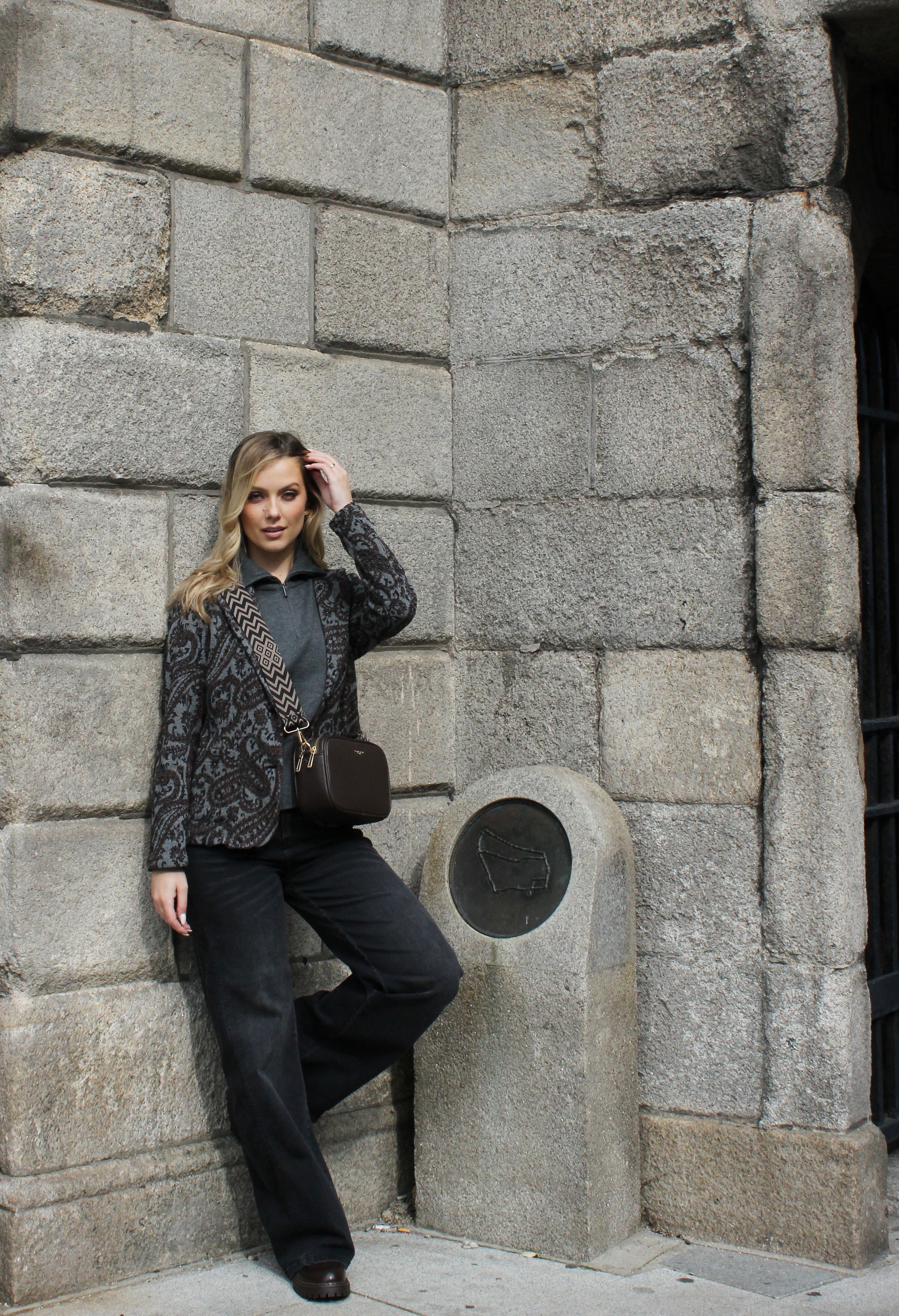 Woman leaning against a stone wall, wearing a patterned jacket and dark pants.