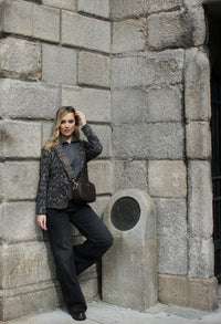 Woman leaning against a stone wall, wearing a patterned jacket and dark pants.