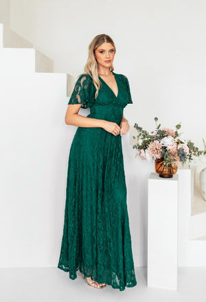 Woman in a green lace dress standing in a minimalistic room with flowers on a table.