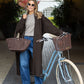 Woman standing next to a light blue bicycle holding a brown bag, wearing a long coat and sunglasses.