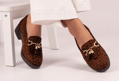 Brown leopard print loafers with gold accents worn by a person sitting on a white chair.