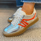 Silver and orange sneakers with white laces worn on a patterned carpet.