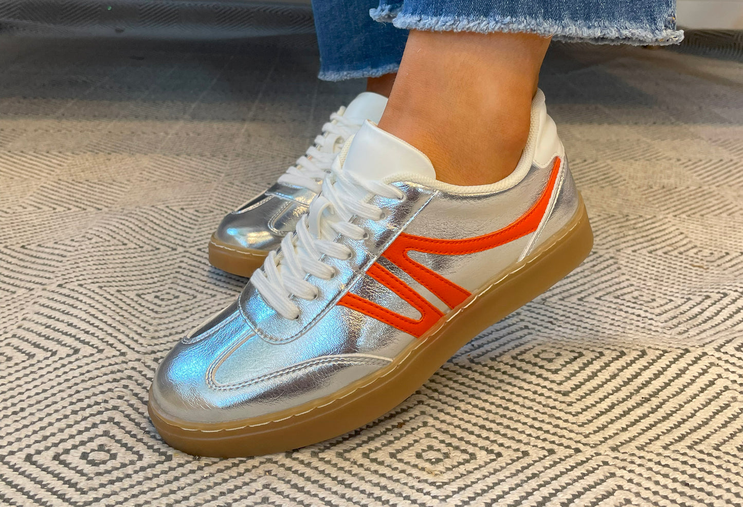 Silver and orange sneakers with white laces worn on a patterned carpet.