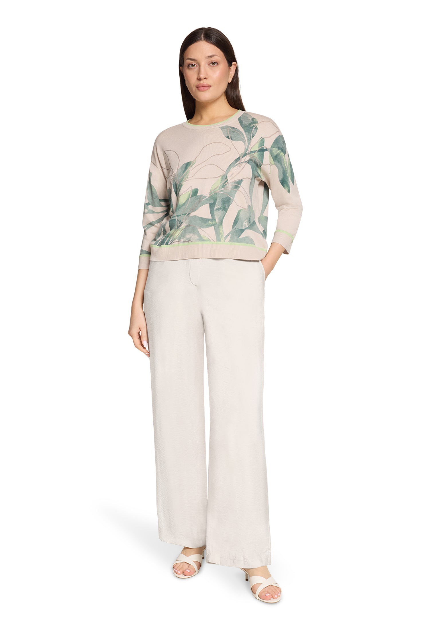 Woman wearing a beige sweater with green leaf pattern and white pants on a white background