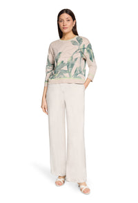 Woman wearing a beige sweater with green leaf pattern and white pants on a white background