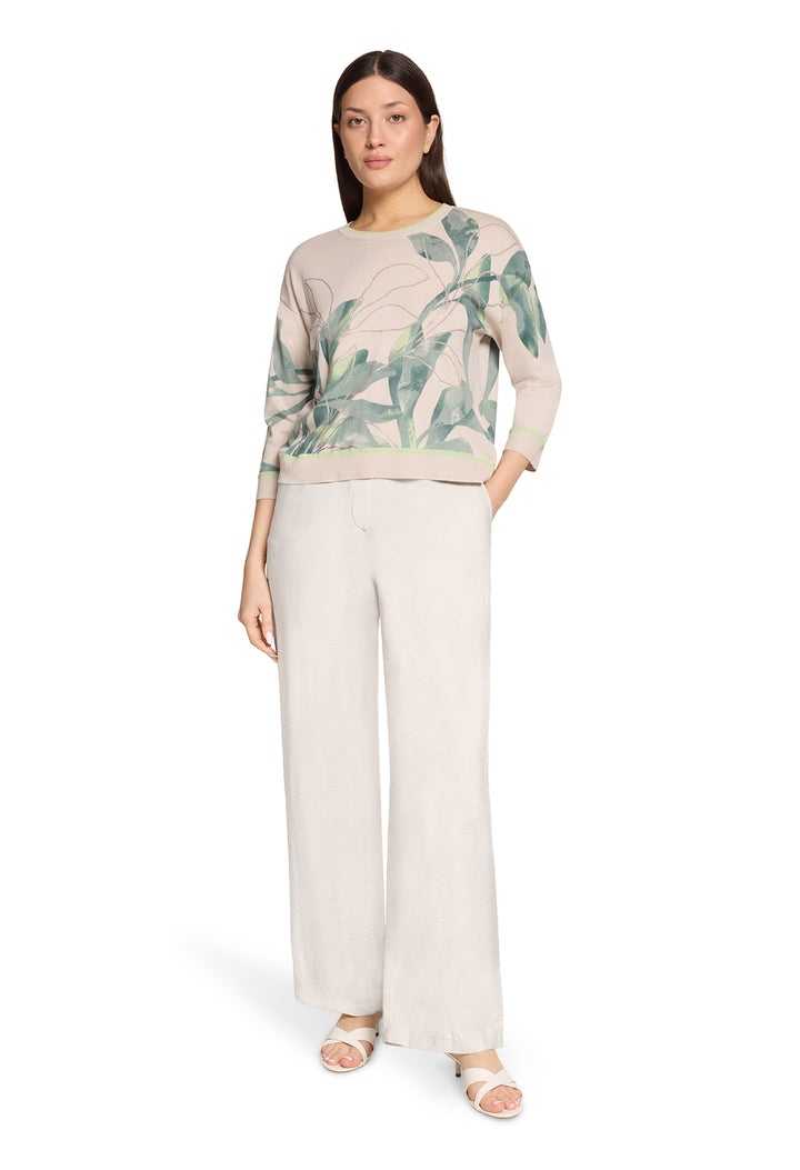 Woman wearing a beige sweater with green leaf pattern and white pants on a white background