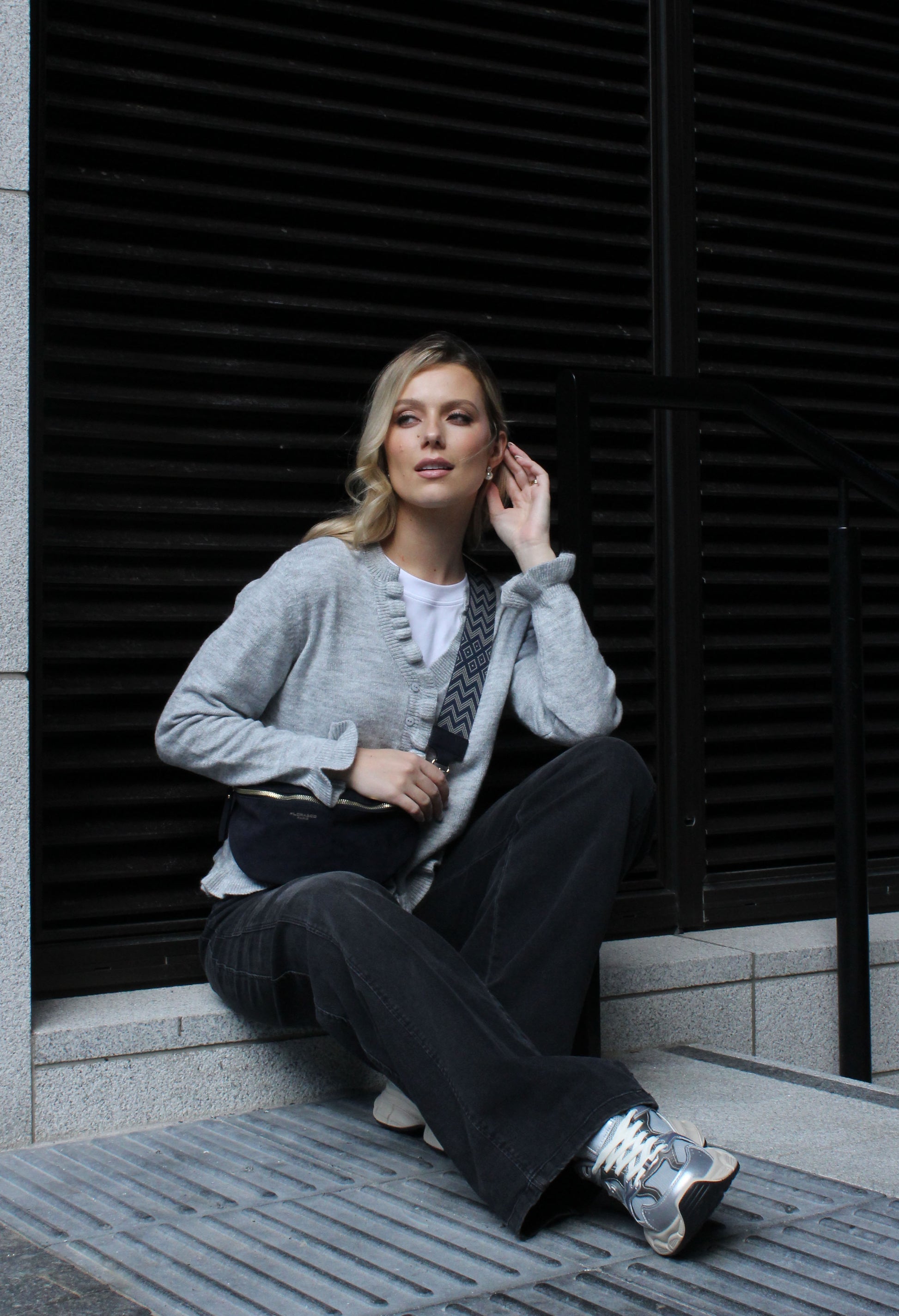 Woman sitting on a step wearing a gray cardigan and dark pants against a black shutter background.