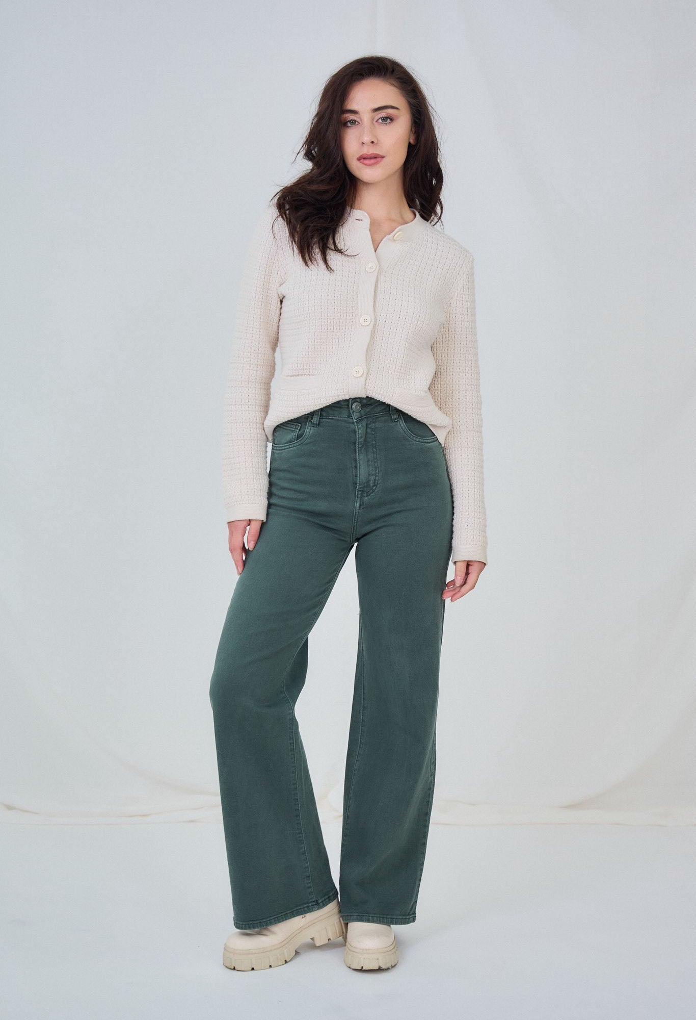 Woman wearing a cream sweater and green pants against a white background