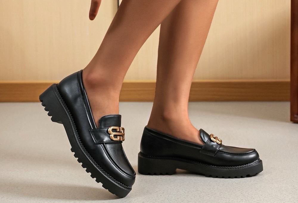 Black loafers with gold buckles worn by a person on a neutral background
